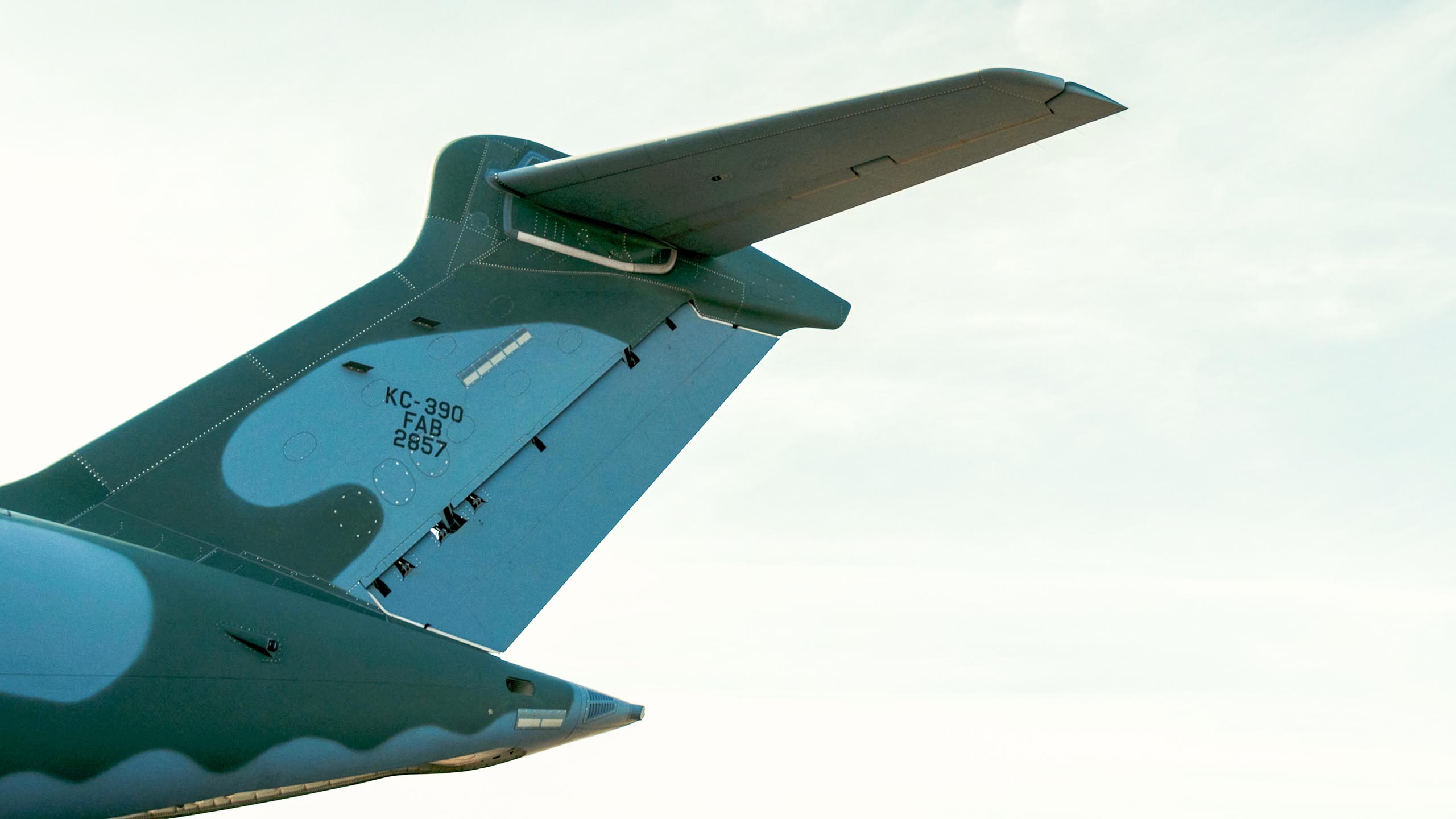 Foto em ângulo baixo (contra-plongée) mostrando a cauda em formato de "T" de um avião militar camuflado contra o céu claro. Na parte vertical da estrutura, lê-se a inscrição "KC-390 FAB 2857" em preto.