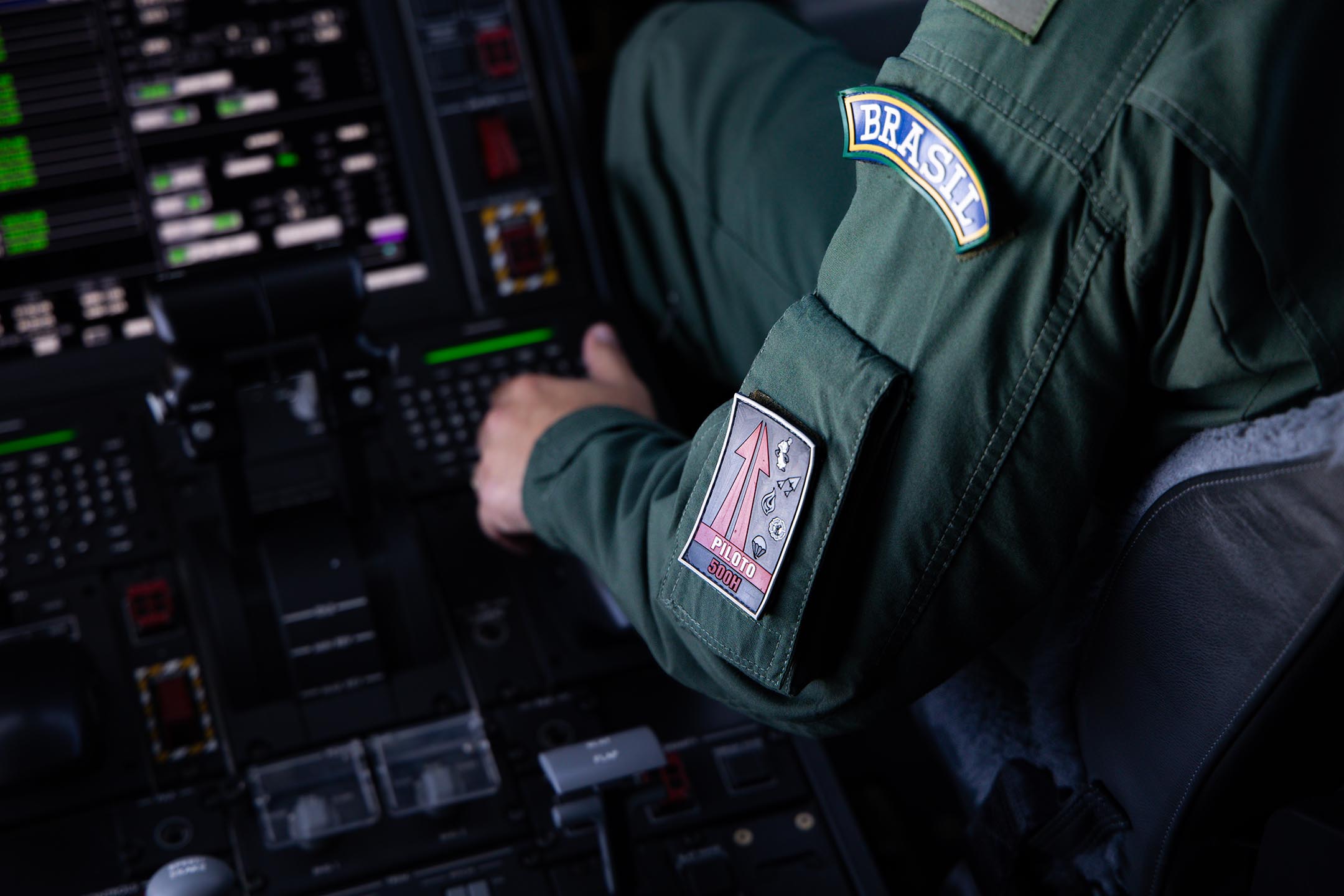 Braço de piloto com macacão verde e insígnia "500H". Ao fundo, luzes do painel desfocadas.